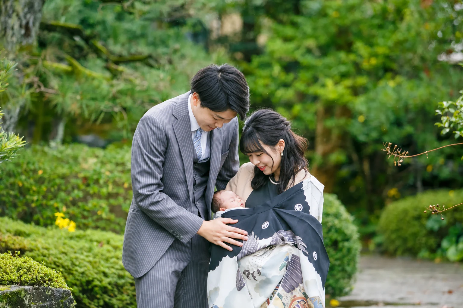 緑豊かな境内で赤ちゃんを見つめるお宮参りの両親